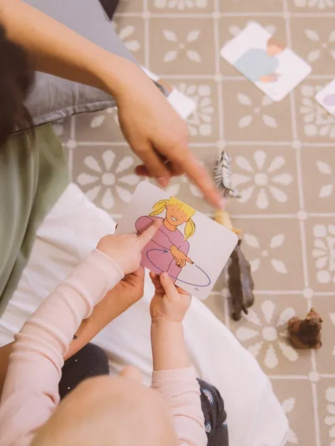 Kinderhände halten eine Bildkarte, die Therapeutin zeigt auf ein Detail