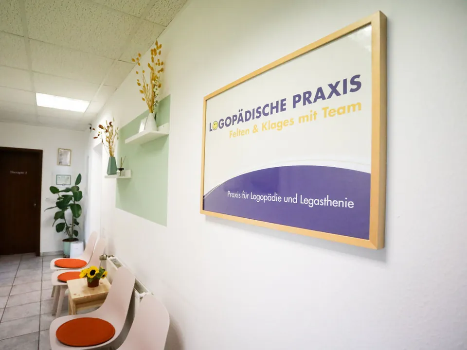 Wartebereich der Praxis Felten & Klages mit gerahmtem Praxisschild „Logopädische Praxis Felten & Klages mit Team“, hellen Stühlen und Sonnenblumen